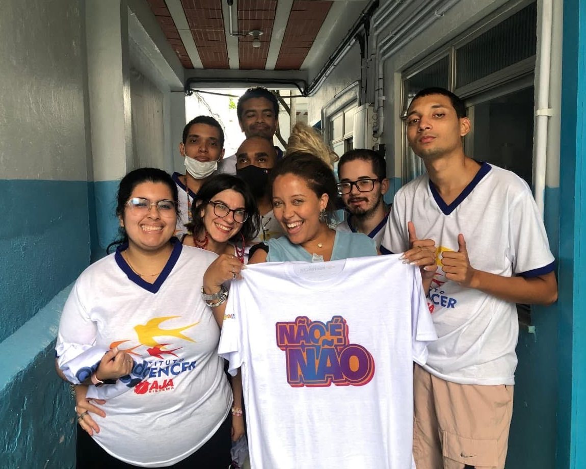 Turma do AJA sorrindo no corredor do Instituto, com camisetas do Sistema AJA, segurando uma camiseta Não é Não