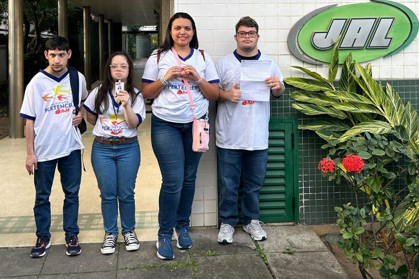 Três participantes do AJA e uma profissional do Instituto, com camisetas do Sistema AJA, em frente à empresa parceira JAL